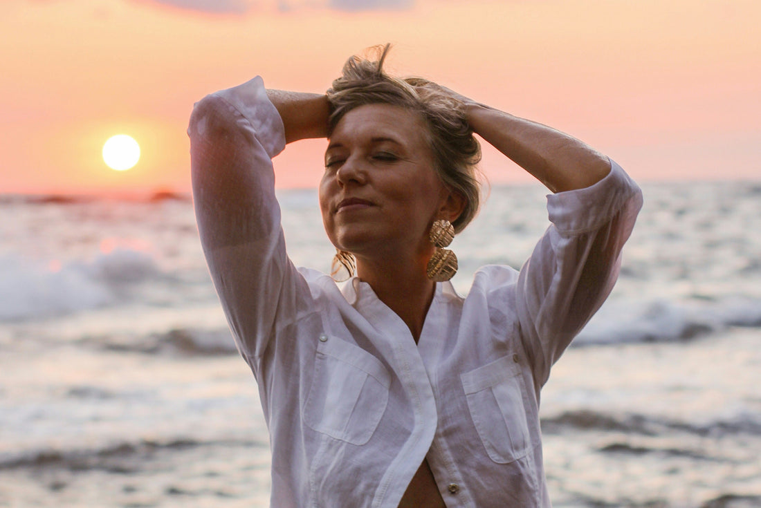 Frau am Meer bei Sonnenuntergang, Hände im Haar, entspannter Ausdruck